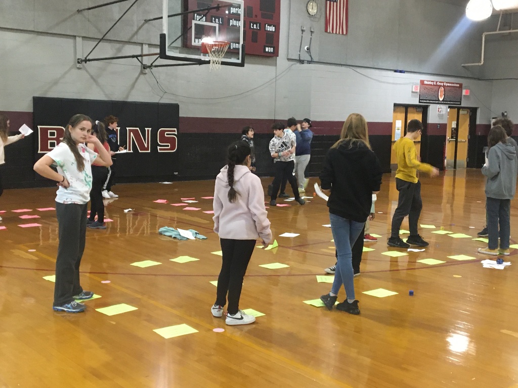Students moving along a board game tiles created by paper on the gym floor